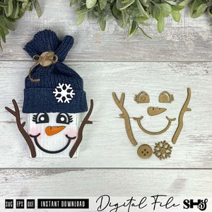 May include: A snowman craft kit featuring a white wooden block with a painted face, a dark blue knit hat with a white snowflake, and two brown twig arms. The kit includes additional wooden cutouts of eyes, a nose, a mouth, arms, and a snowflake.