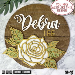 May include: A circular wooden sign featuring a dark brown stained plank background. The name Debra is written in white script, with the word LEE in gold block letters below. A layered floral design in gold and light green sits at the bottom.