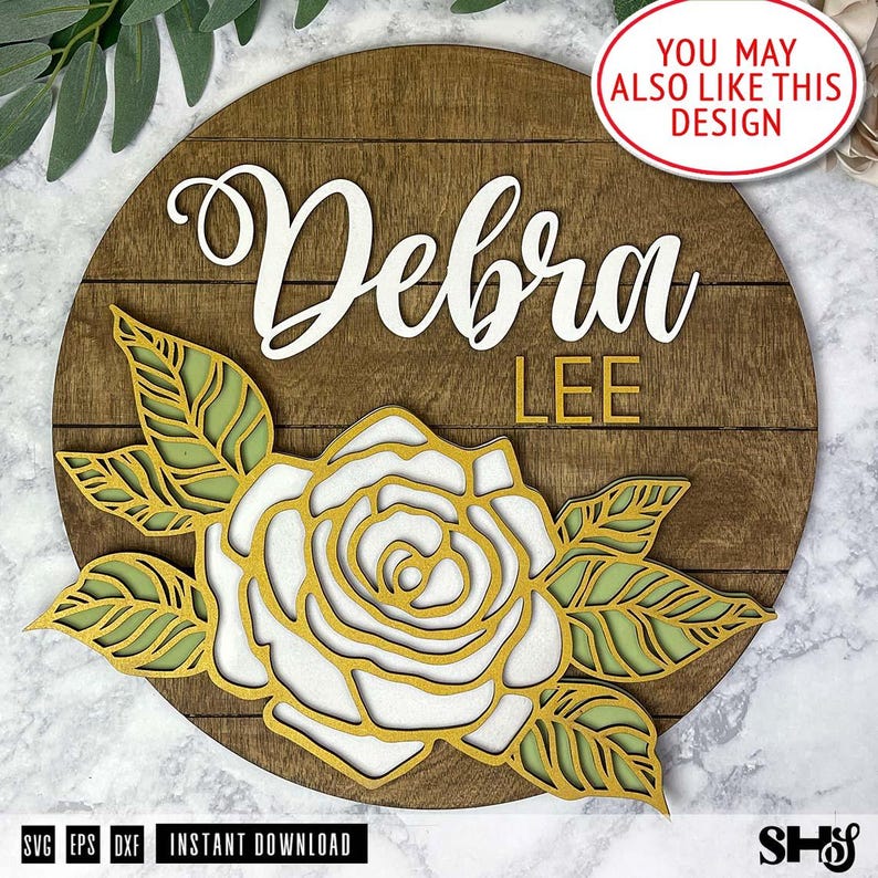 May include: A circular wooden sign featuring a dark brown stained plank background. The name Debra is written in white script, with the word LEE in gold block letters below. A layered floral design in gold and light green sits at the bottom.