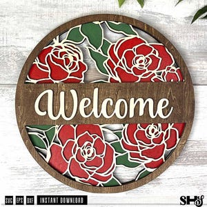May include: A circular wooden sign featuring a layered design with red roses and green leaves. A horizontal wooden bar across the centre displays the word Welcome in a cream-coloured script font. The piece has a dark brown stained wood frame.