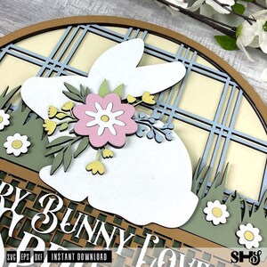 Pode incluir: Uma placa circular de madeira em camadas com uma silhueta de coelho branco adornada com uma flor rosa e folhas verdes. O fundo apresenta um padr&atilde;o xadrez azul claro e creme, grama verde com flores brancas e o texto "BUNNY LOVE".