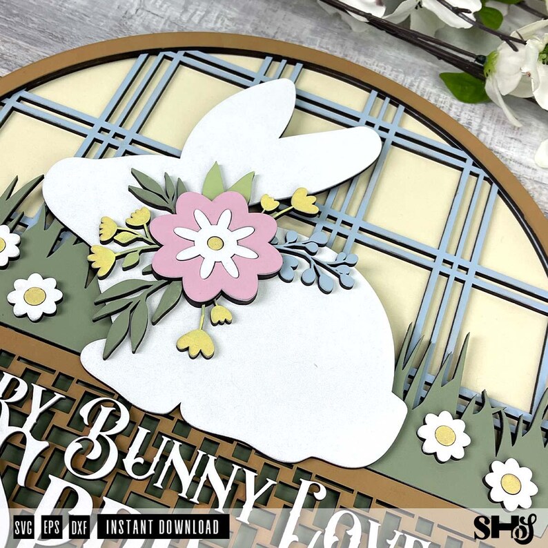 Pode incluir: Uma placa circular de madeira em camadas com uma silhueta de coelho branco adornada com uma flor rosa e folhas verdes. O fundo apresenta um padr&atilde;o xadrez azul claro e creme, grama verde com flores brancas e o texto "BUNNY LOVE".