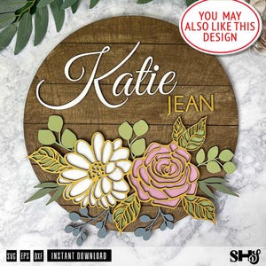May include: A circular wooden sign featuring the name Katie Jean in white and yellow script and block lettering. The design includes layered floral cutouts in white, pink, yellow, and green, with various leaf shapes and stems arranged below.
