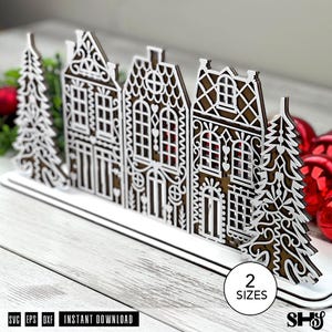 May include: A laser-cut wooden decorative display featuring a row of three intricate houses flanked by two evergreen trees. The design includes white filigree overlays on a brown base. A circular white label displays the text 2 SIZES.