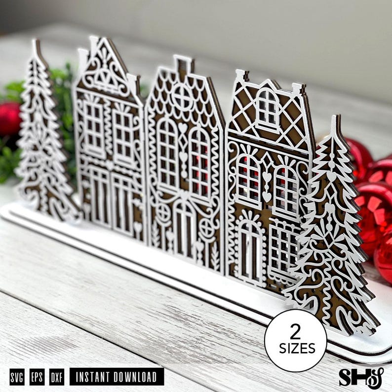 May include: A laser-cut wooden decorative display featuring a row of three intricate houses flanked by two evergreen trees. The design includes white filigree overlays on a brown base. A circular white label displays the text 2 SIZES.