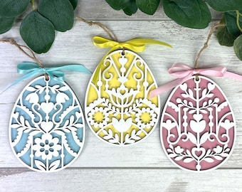 May include: Three Easter egg ornaments in blue, yellow, and pink, each with a white, intricate floral design. The eggs are layered, with a contrasting color visible through the cut-out patterns. Each egg has a ribbon and twine for hanging.