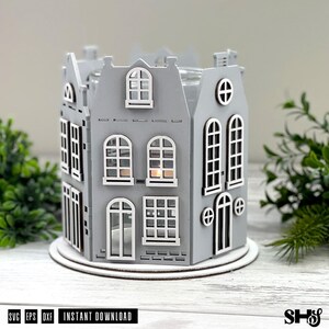 May include: A gray laser-cut wooden candle holder shaped like a cluster of houses with white-trimmed windows and doors. The structure features multiple arched and rectangular window cutouts, a central light source, and a circular base. Text reads SVG EPS DXF.