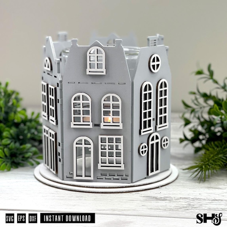 May include: A gray laser-cut wooden candle holder shaped like a cluster of houses with white-trimmed windows and doors. The structure features multiple arched and rectangular window cutouts, a central light source, and a circular base. Text reads SVG EPS DXF.