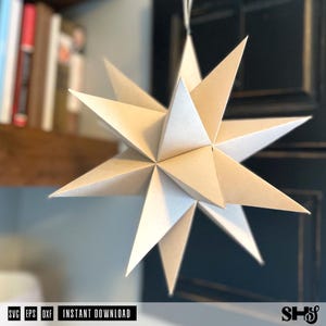 May include: A three-dimensional paper star ornament featuring multiple pointed triangular segments arranged in a geometric pattern. The star is off-white in color and hangs from a thin string, displaying sharp edges and symmetrical facets.
