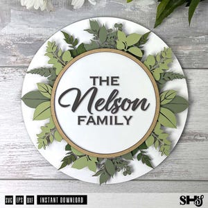 May include: A circular white wall sign featuring a central wooden hoop frame. The text "THE Nelson FAMILY" is displayed in the center. The sign is decorated with layered, die-cut paper or wood leaves in various shades of green and sage.