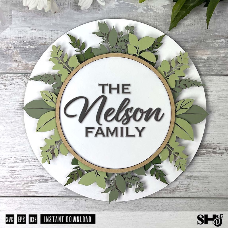 May include: A circular white wall sign featuring a central wooden hoop frame. The text "THE Nelson FAMILY" is displayed in the center. The sign is decorated with layered, die-cut paper or wood leaves in various shades of green and sage.