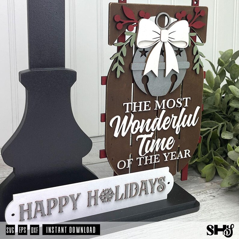 May include: A layered wooden sled decoration featuring a grey bell with a white bow, red berries, and green leaves. The text reads "THE MOST WONDERFUL TIME OF THE YEAR". A separate white sign in front displays the text "HAPPY HOLIDAYS" with a snowflake.