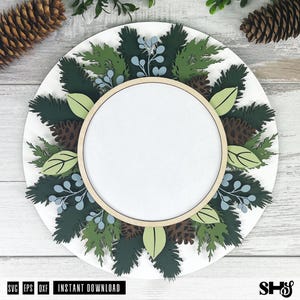 May include: A circular layered sign featuring a central blank white space framed by a light wood hoop. The design includes dark green pine branches, light green leaves, brown pinecones, and light blue berries arranged in a wreath pattern.