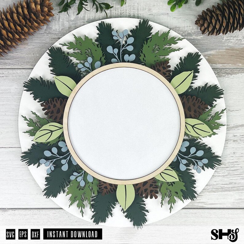 May include: A circular layered sign featuring a central blank white space framed by a light wood hoop. The design includes dark green pine branches, light green leaves, brown pinecones, and light blue berries arranged in a wreath pattern.