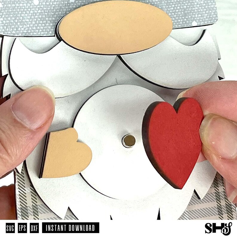 May include: A layered wooden gnome craft project featuring a light grey textured hat, a tan oval nose, a white beard, and a central circular piece with a small metal pin. A small tan heart and a larger red heart are positioned near the centre.