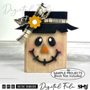 Puede incluir: Un bloque de madera de espantapájaros con un sombrero negro, una flor amarilla y una cinta a cuadros. El espantapájaros tiene ojos negros, una nariz de madera naranja y una boca sonriente. El texto "SAMPLE PROJECTS Block Not Included" es visible.