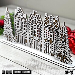 May include: A decorative laser-cut wooden display featuring a row of three intricate gingerbread-style houses flanked by two stylized evergreen trees. The design features white filigree overlays on a brown base, mounted on a flat white stand.