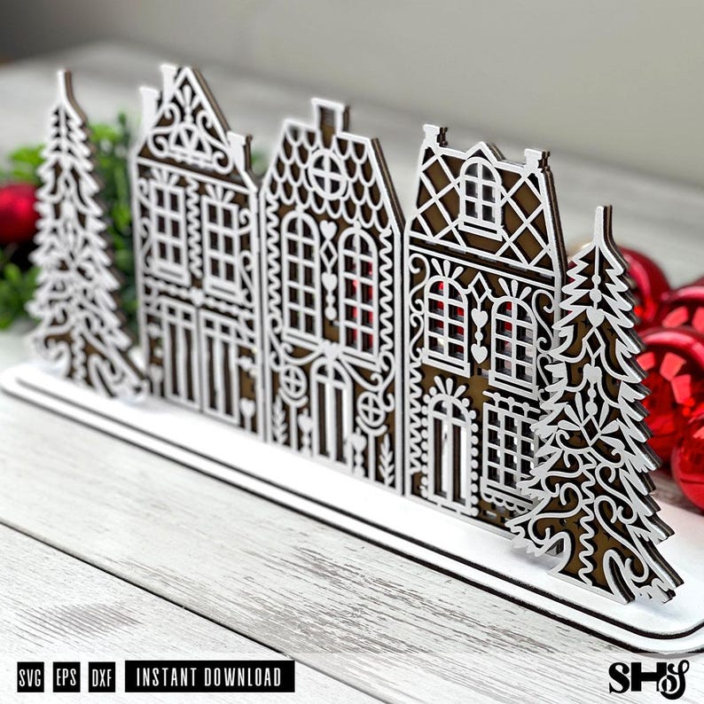 May include: A decorative laser-cut wooden display featuring a row of three intricate gingerbread-style houses flanked by two stylized evergreen trees. The design features white filigree overlays on a brown base, mounted on a flat white stand.