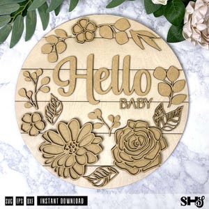 May include: A circular light-colored wooden sign featuring the laser-cut text Hello BABY in the center. The sign is decorated with layered wooden floral and leaf cutouts arranged around the text, including large flowers and sprigs of foliage.