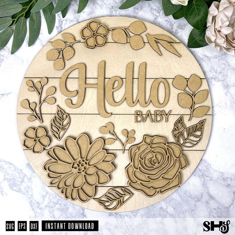 May include: A circular light-colored wooden sign featuring the laser-cut text Hello BABY in the center. The sign is decorated with layered wooden floral and leaf cutouts arranged around the text, including large flowers and sprigs of foliage.