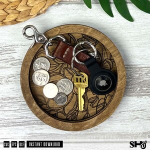 May include: A round wooden tray with a laser-engraved floral pattern containing a brown leather keychain with a silver metal clasp, a gold-colored key, a black circular tracking device, and four silver-colored coins resting on the tray surface.