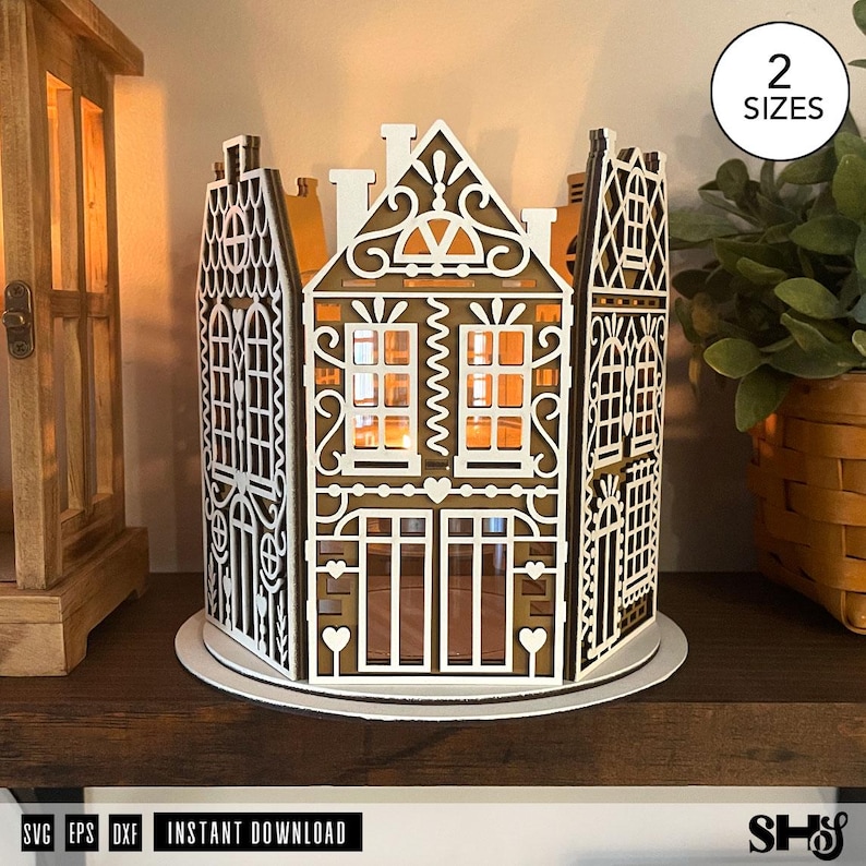 May include: A laser-cut wooden gingerbread house luminary featuring intricate white filigree patterns, arched windows, and heart motifs. The structure is composed of multiple panels arranged in a circular formation on a round base, with the text 2 SIZES.