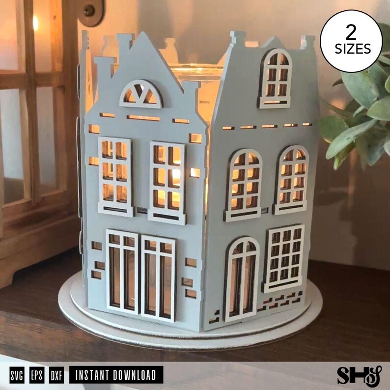 May include: A light gray laser-cut wooden house-shaped lantern featuring multiple windows and a gabled roof design. The structure is illuminated from within, casting a warm glow through the cutouts. A circular white base supports the house.