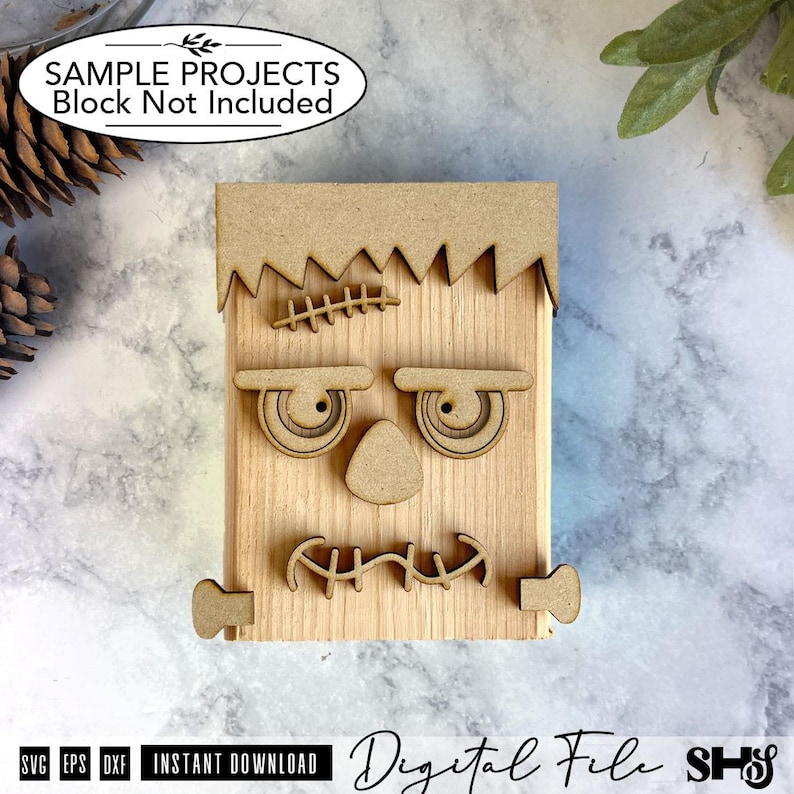 May include: A light-colored wooden block decorated with laser-cut wooden pieces forming a face. The design includes a jagged hairline, a stitched forehead scar, two eyes, a triangular nose, a stitched mouth, and two small rectangular side bolts.