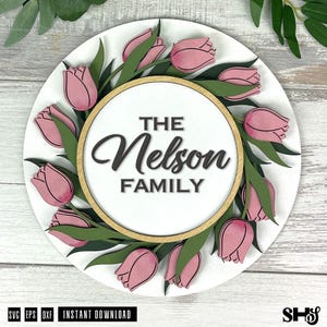 May include: A circular wooden sign featuring a layered design of pink tulips and green leaves surrounding a central white circle. The text THE Nelson FAMILY is printed in black in the center. The sign has a light wood-toned circular frame.