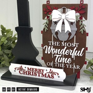 May include: A brown wooden sled-shaped sign featuring a grey jingle bell with a white bow, decorated with red and green foliage. The text reads THE MOST WONDERFUL TIME OF THE YEAR. A separate white sign base displays the text MERRY CHRISTMAS in red.