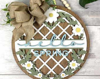 May include: A wooden round sign with a white lattice design and the words "hello spring" in blue lettering. The sign is decorated with a brown burlap bow, green leaves, and white flowers.