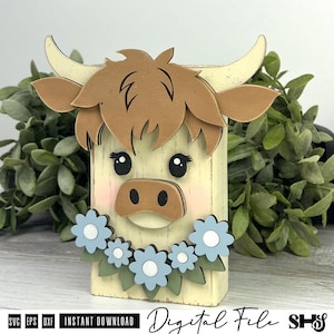 Pu&ograve; includere: Un oggetto artigianale in legno a forma di muso di mucca Highland stilizzato, con pelo marrone chiaro, corna color crema e muso marrone chiaro. La mucca ha occhi neri, guance rosa e una ghirlanda di fiori azzurri con centri bianchi.