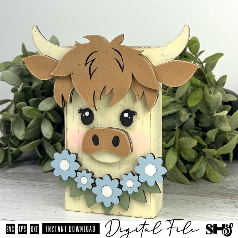 Pu&ograve; includere: Un oggetto artigianale in legno a forma di muso di mucca Highland stilizzato, con pelo marrone chiaro, corna color crema e muso marrone chiaro. La mucca ha occhi neri, guance rosa e una ghirlanda di fiori azzurri con centri bianchi.