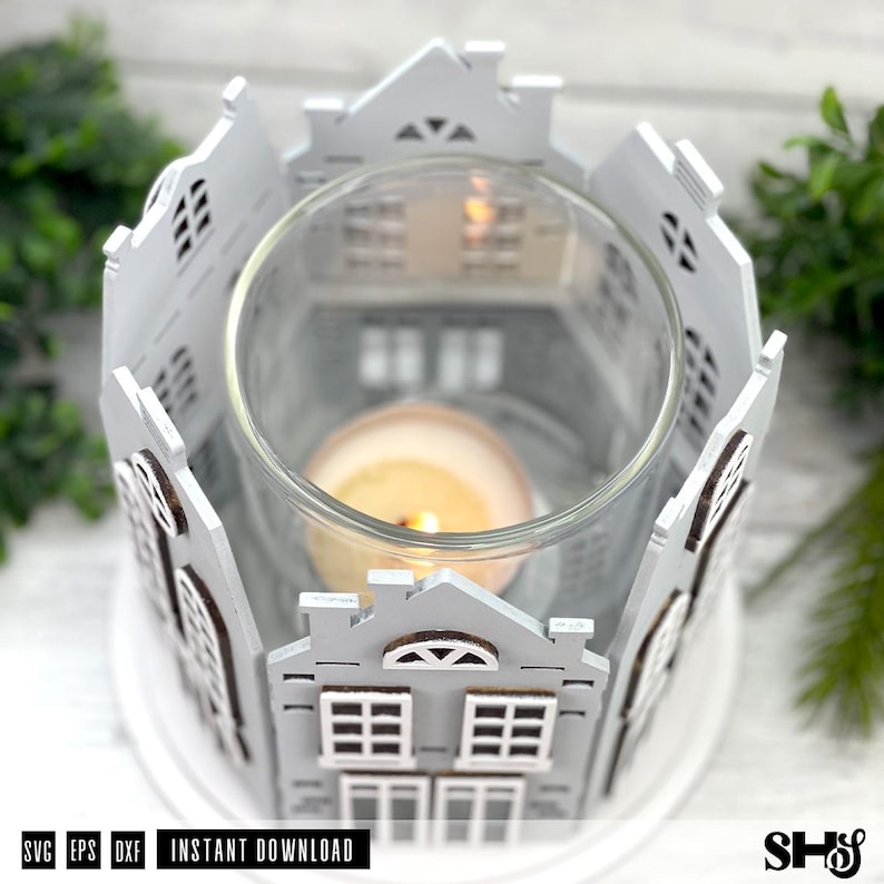 May include: A circular candle holder featuring a white wooden facade with cut-out house designs surrounding a clear glass cylinder. A single lit tea light candle sits inside the glass, casting a warm glow through the windows of the miniature houses.