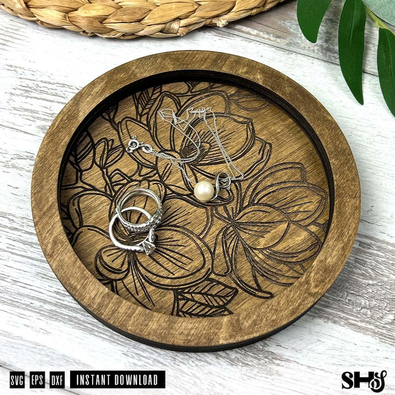 May include: A round wooden tray with a dark brown finish featuring a laser-engraved floral pattern on the base. Inside the tray, there is a silver-toned necklace with a single white pearl pendant and two silver-toned rings stacked together.