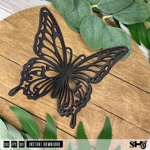 May include: A dark brown, laser-cut butterfly silhouette featuring intricate, open-work wing patterns. The butterfly is positioned against a light brown wooden surface with visible grain, surrounded by several green eucalyptus leaves.