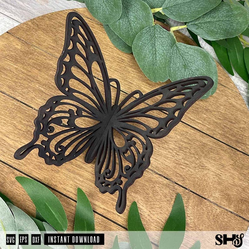 May include: A dark brown, laser-cut butterfly silhouette featuring intricate, open-work wing patterns. The butterfly is positioned against a light brown wooden surface with visible grain, surrounded by several green eucalyptus leaves.