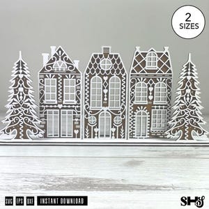May include: A digital file set featuring a brown and white gingerbread village scene with three houses and two pine trees. The design includes intricate white icing-style patterns on the brown structures. A circle in the corner reads 2 SIZES.