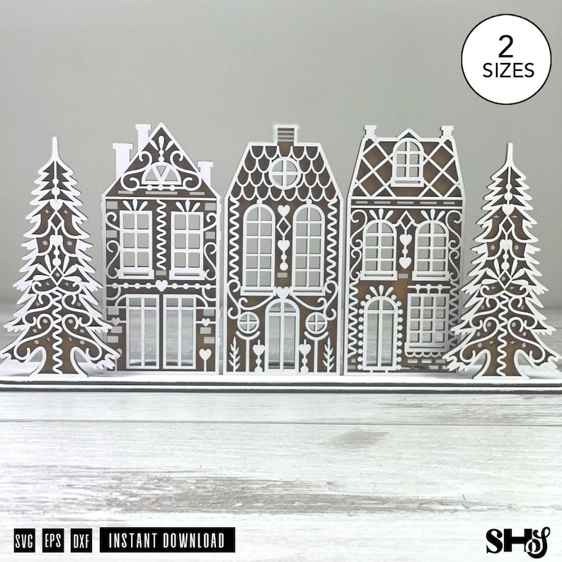 May include: A digital file set featuring a brown and white gingerbread village scene with three houses and two pine trees. The design includes intricate white icing-style patterns on the brown structures. A circle in the corner reads 2 SIZES.