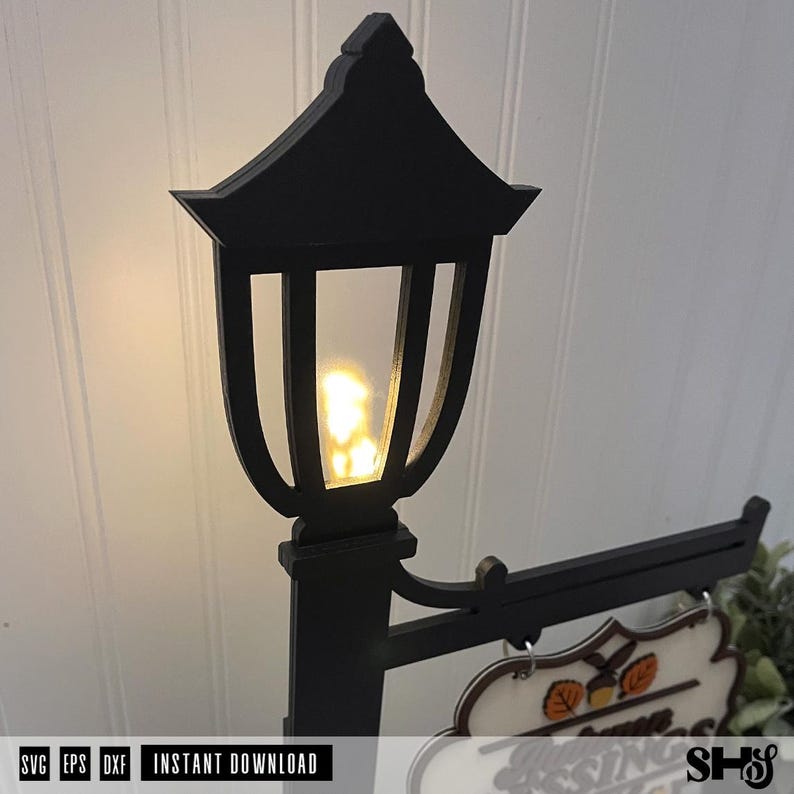 Op de afbeelding: Een zwarte decoratieve lantaarnpaal-uitsparing met een gloeiend geel lichteffect in de lantaarnkop. Aan de zijkant bevindt zich een horizontale arm voor het ophangen van een bord, met een gedeeltelijk zicht op een bord met herfstbladmotieven.