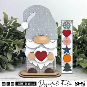 May include: A layered wooden gnome figurine featuring a grey polka-dot hat, a white beard, and a tan nose. The gnome holds a red heart. Beside it is a vertical wooden strip with interchangeable icons: a snowflake, heart, flower, star, pumpkin, and tree.