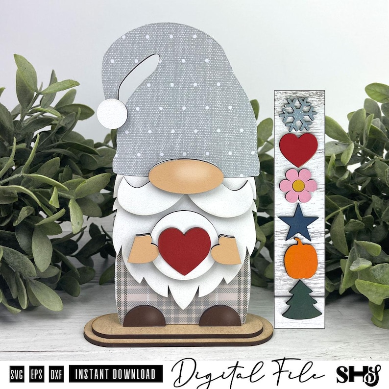 May include: A layered wooden gnome figurine featuring a grey polka-dot hat, a white beard, and a tan nose. The gnome holds a red heart. Beside it is a vertical wooden strip with interchangeable icons: a snowflake, heart, flower, star, pumpkin, and tree.