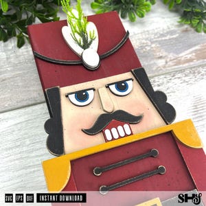 May include: A wooden nutcracker decoration featuring a red hat with a white plume, black hair, blue eyes, a black moustache, and a red jacket with yellow accents. The design includes two horizontal black lines on the chest and white teeth.