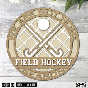 Może przedstawiać: Okrągła drewniana tabliczka z tłem w kratkę, dwoma skrzyżowanymi kijami do hokeja na trawie i piłką. Napis WE ARE NOT HOME FIELD HOCKEY SEASON jest wycięty w drewnie. Projekt utrzymany jest w jasnobrązowej i białej kolorystyce.