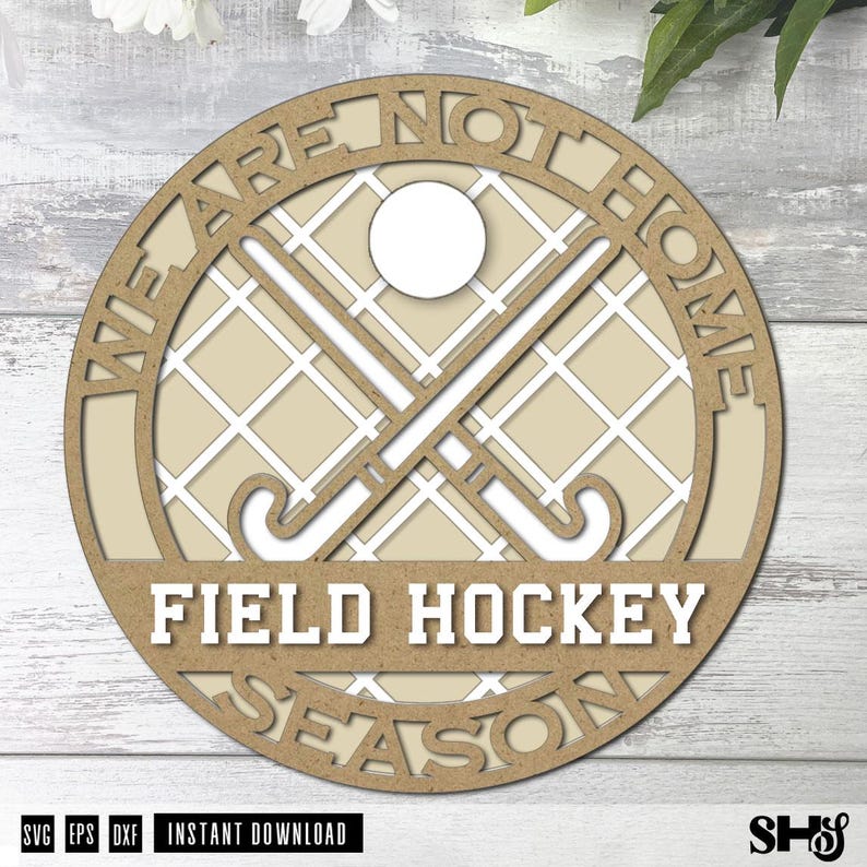 Może przedstawiać: Okrągła drewniana tabliczka z tłem w kratkę, dwoma skrzyżowanymi kijami do hokeja na trawie i piłką. Napis WE ARE NOT HOME FIELD HOCKEY SEASON jest wycięty w drewnie. Projekt utrzymany jest w jasnobrązowej i białej kolorystyce.