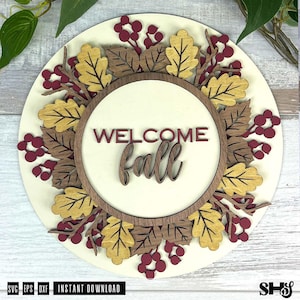 May include: A circular wooden sign featuring the text "WELCOME fall" in dark red lettering. The sign is decorated with a layered wreath of laser-cut wooden oak leaves in shades of yellow and brown, interspersed with clusters of dark red berries.