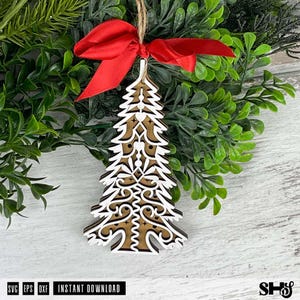 Puede incluir: Adorno de &aacute;rbol de Navidad de madera con intrincados dise&ntilde;os calados blancos y un fondo marr&oacute;n. Est&aacute; adornado con un lazo de sat&eacute;n rojo y una cuerda para colgar. El adorno se coloca sobre un fondo de follaje verde.