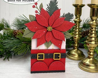 May include: A decorative craft project featuring a large red poinsettia flower with green leaves and pine sprigs atop a red base resembling Santa Claus boots with black belts and gold buckles. The text reads SAMPLE PROJECTS Block not included.