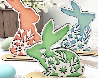 May include: Three wooden bunny silhouettes with floral designs. The bunnies are painted in shades of pink, blue, and green. The bunnies are standing on wooden bases.