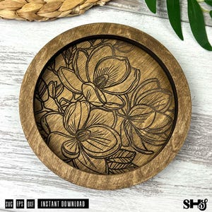 May include: A circular wooden coaster featuring a laser-engraved floral design of magnolia blossoms with detailed petals and leaves. The natural wood grain is visible throughout the piece, which has a raised outer rim and a recessed center.